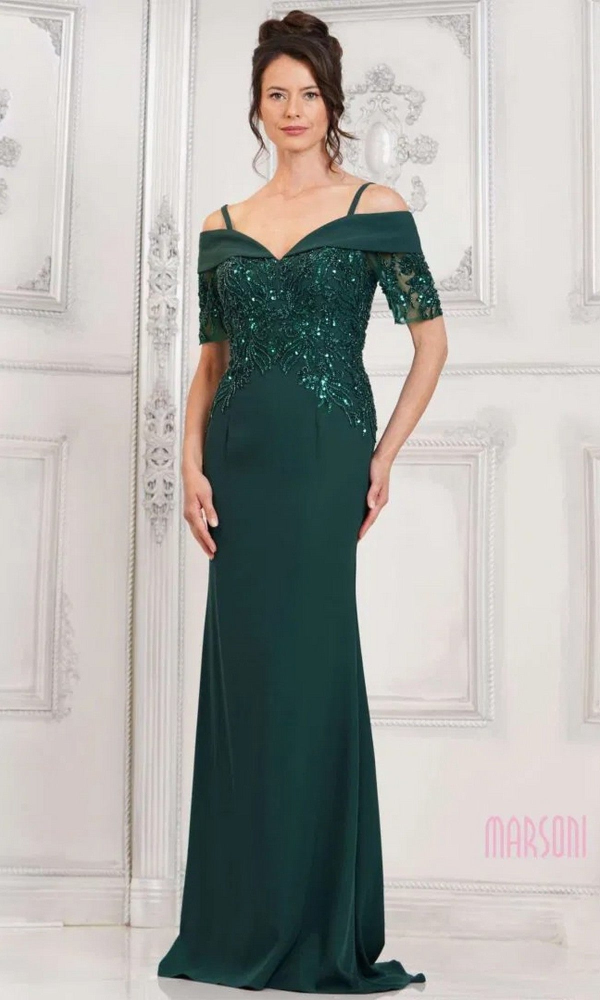 A woman in the Marsoni MV1295 long formal dress in deep green, featuring sequin appliques, off-the-shoulder sleeves, and spaghetti straps, stands against a white ornate background.